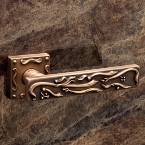 Könnte beinhalten: Ein bronzener Türgriff mit einem verzierten Design mit einem floralen Motiv. Der Griff hat eine quadratische Rückplatte mit dekorativen Akzenten.