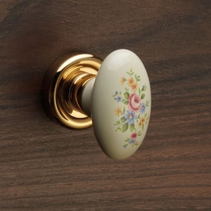 Può includere: Una manopola per porta in ceramica bianca con un design floreale in tonalità rosa, giallo e blu. La manopola è di forma ovale e ha una base in metallo dorato.