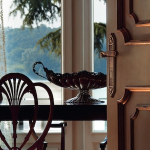Könnte beinhalten: Eine silberne Servierschale mit einem verzierten Design steht auf einem dunklen Holztisch vor einem Fenster mit Blick auf einen See und Bäume. Zwei Holzstühle mit komplizierten Designs sind im Vordergrund zu sehen.
