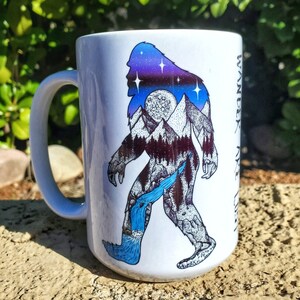 Pu&ograve; includere: Tazza in ceramica bianca con la silhouette di un Bigfoot, riempita con una scena di montagna e cascata sotto un cielo stellato. La tazza ha un grande manico e la parola "Wander" &egrave; stampata verticalmente sul lato.