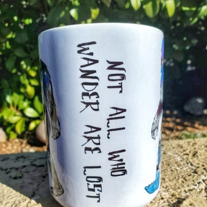 Pu&ograve; includere: Tazza in ceramica bianca con la scritta "NOT ALL WHO WANDER ARE LOST" in nero. La tazza presenta illustrazioni di un lupo e altri disegni colorati. La tazza &egrave; posta su uno sfondo naturale.