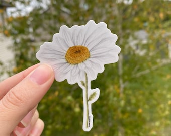 May include: A white daisy sticker with a yellow center and green stem. The sticker is on a white background and has a handwritten signature at the bottom.