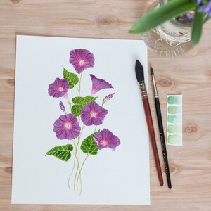 May include: A watercolor painting of purple morning glory flowers with green leaves on a white background. Two paintbrushes and a watercolor palette are visible on a wooden surface.