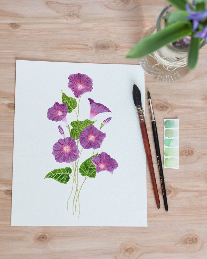 May include: A watercolor painting of purple morning glory flowers with green leaves on a white background. Two paintbrushes and a watercolor palette are visible on a wooden surface.