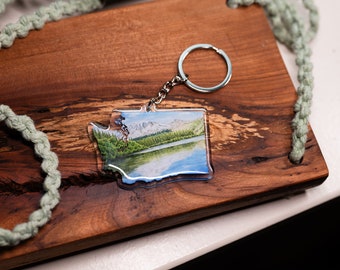 Puede incluir: Un llavero de acrílico transparente con forma del estado de Washington con una imagen escénica de un lago y montañas en su interior. El llavero tiene un anillo de metal plateado.