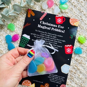 Peut inclure: Un petit sac en filet transparent rempli de galets colorés et lisses. Le sac est tenu dans une main avec des ongles aux couleurs de l'arc-en-ciel, à côté d'une carte noire avec le texte "Christmas Eve Magical Pebbles!". D'autres galets brillants sont éparpillés.