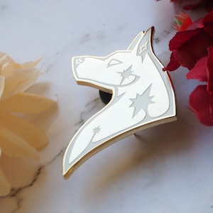 Puede incluir: Pin de esmalte blanco de una cabeza de lobo con contorno dorado y detalles de estrellas blancas.