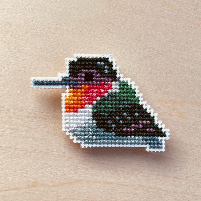 Op de afbeelding: Een kruissteek kolibrie speld met een witte rand. De vogel heeft een zwarte kop, rode keel, oranje snavel en groen en blauw lichaam. De speld is geplaatst tegen een lichte houtnerf achtergrond.