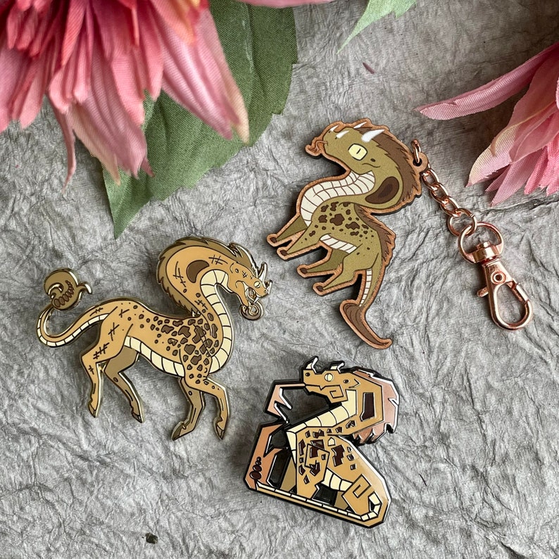 Op de afbeelding: Drie gouden emaille-speldjes met verschillende mythische wezens. De eerste speld is een bruine en gouden draak met een lange nek en staart. De tweede speld is een bruine en gouden slang met een lange staart. De derde speld is een bruine en gouden draak met een lange staart en een korte nek.