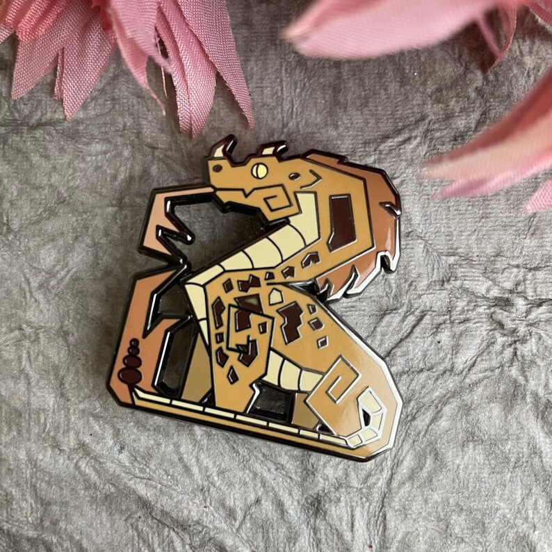 Op de afbeelding: Een bruine en beige emaille pin van een draak met een geometrisch ontwerp. De draak is opgerold met zijn kop naar de rechterkant van de pin gericht.