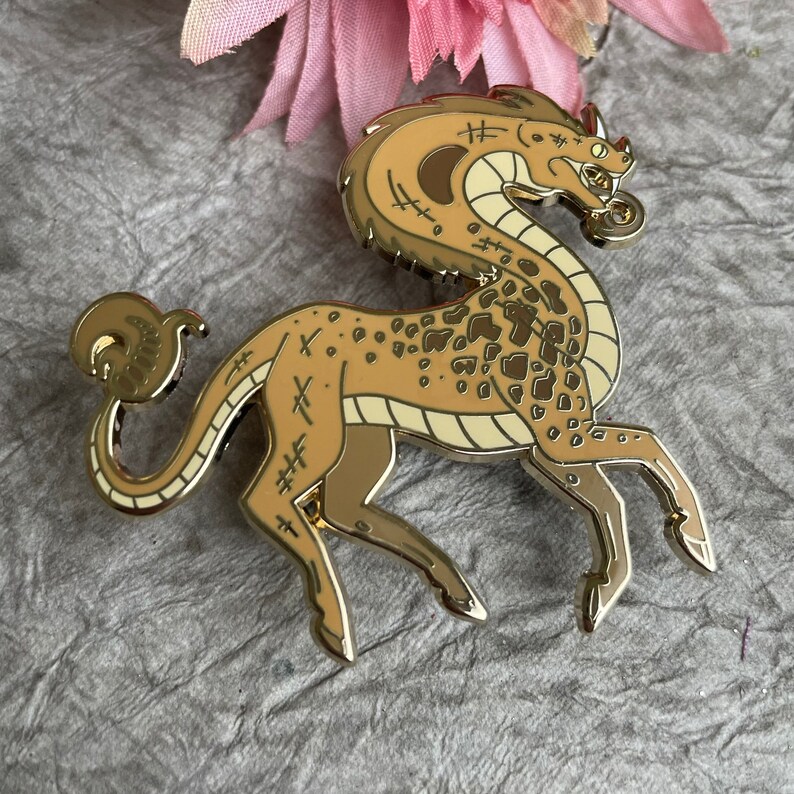 Op de afbeelding: Gouden emaille pin van een mythisch wezen met een paardenlichaam, een lange nek en een drakenkop. Het wezen heeft een gevlekt patroon en is versierd met stiksels.