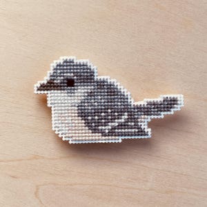 Op de afbeelding: Een kruissteekvogel in tinten grijs, zwart en wit, met een witte rand. De vogel is geplaatst op een lichtbruin houten oppervlak. Het ontwerp van de vogel heeft een gedetailleerd patroon dat de textuur van de kruissteek laat zien.