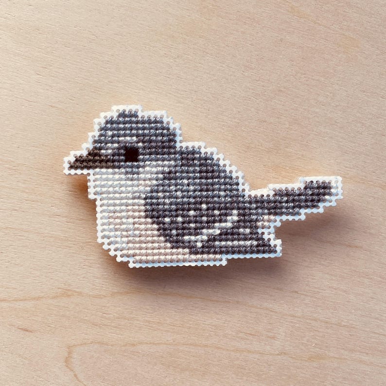 Op de afbeelding: Een kruissteekvogel in tinten grijs, zwart en wit, met een witte rand. De vogel is geplaatst op een lichtbruin houten oppervlak. Het ontwerp van de vogel heeft een gedetailleerd patroon dat de textuur van de kruissteek laat zien.