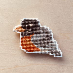 Op de afbeelding: Een kruissteek vogelspeld met een roodborstje ontwerp. De vogel heeft een zwarte kop, een grijze en bruine body en een fel oranje borst. De speld is wit omlijnd en geplaatst tegen een lichte houtnerf achtergrond.