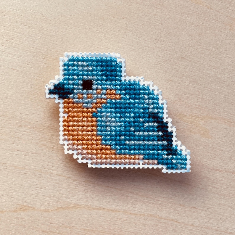Op de afbeelding: Een kruissteekvogel in blauw- en oranje tinten, met een witte rand. De vogel wordt afgebeeld in een zittende positie, met zijn vleugels gevouwen. De achtergrond is een lichte houtnerf.