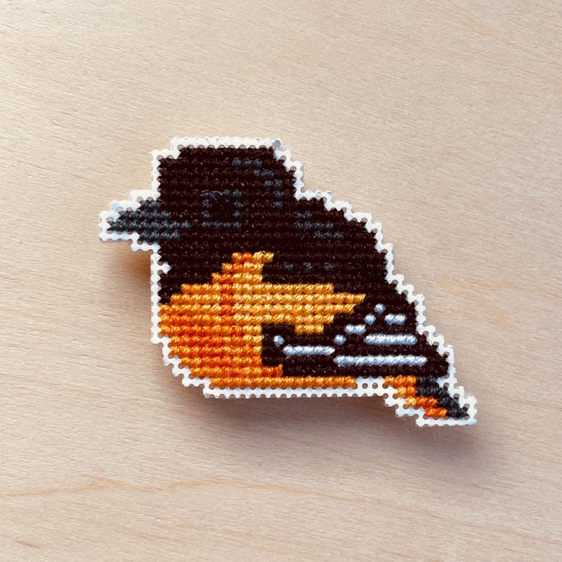 Op de afbeelding: Een kruissteekvogel in zwart, oranje en wit. De vogel heeft een zwarte kop en rug, een oranje borst en buik en witte accenten. De vogel staat op een licht houten oppervlak.