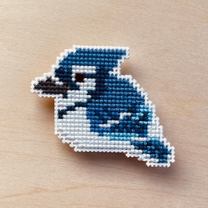 Op de afbeelding: Een kruissteekontwerp in de vorm van een blauwe gaai. De vogel is weergegeven in tinten blauw, wit en zwart, met een getextureerde afwerking. Het handwerk staat tegen een lichte houten achtergrond, wat de handgemaakte aard van het item benadrukt.
