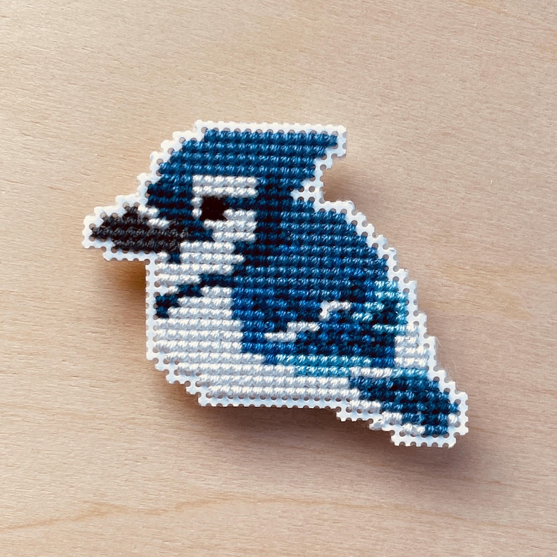 Op de afbeelding: Een kruissteekontwerp in de vorm van een blauwe gaai. De vogel is weergegeven in tinten blauw, wit en zwart, met een getextureerde afwerking. Het handwerk staat tegen een lichte houten achtergrond, wat de handgemaakte aard van het item benadrukt.