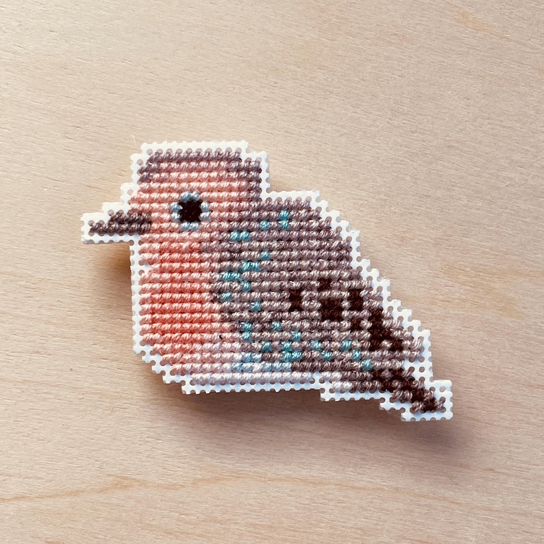 Op de afbeelding: Een kruissteek vogelspeld met een witte rand. De vogel heeft een rode borst, een bruin en blauw lichaam en een zwart oog. De speld staat tegen een lichte houtnerf achtergrond.