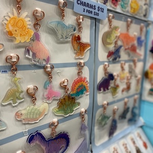 May include: A selection of dinosaur keyrings with rose gold fixings. The keyrings showcase diverse, colourful dinosaur designs, such as a triceratops, a blue T-Rex, and a purple dinosaur. A sign displays the price of the charms.