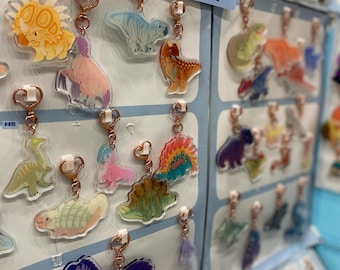 May include: A display of dinosaur-themed keychains with rose gold hardware. The keychains feature various colorful dinosaur designs, including a triceratops, a blue T-Rex, and a purple dinosaur. A sign indicates the price of the charms.