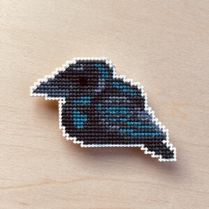 Op de afbeelding: Een kruissteek raaf met een witte rand. De vogel is voornamelijk zwart met teal en grijze accenten, tegen een lichte houtnerf achtergrond. De raaf is een decoratief item.
