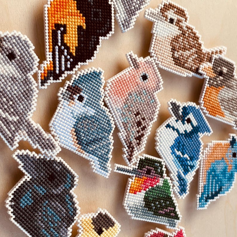 Op de afbeelding: Een verzameling kleurrijke, kruisgestikte vogelornamenten. De vogels hebben verschillende ontwerpen, waaronder een kolibrie, een blauwe gaai en andere soorten, met witte randen. De ornamenten zijn geplaatst tegen een lichtbruine achtergrond.