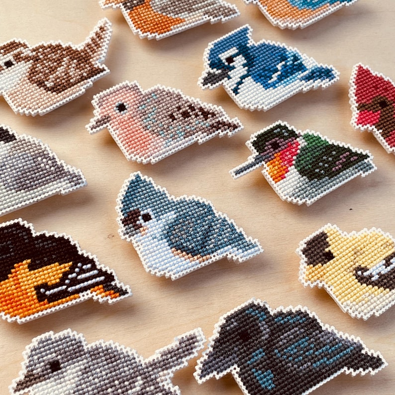 Op de afbeelding: Een verzameling kleurrijke, kruissteek vogelornamenten. De vogels tonen verschillende soorten, waaronder een blauwe gaai en een kolibrie, met gedetailleerde patronen en witte randen. Deze decoratieve stukken zijn gerangschikt op een licht houten oppervlak.