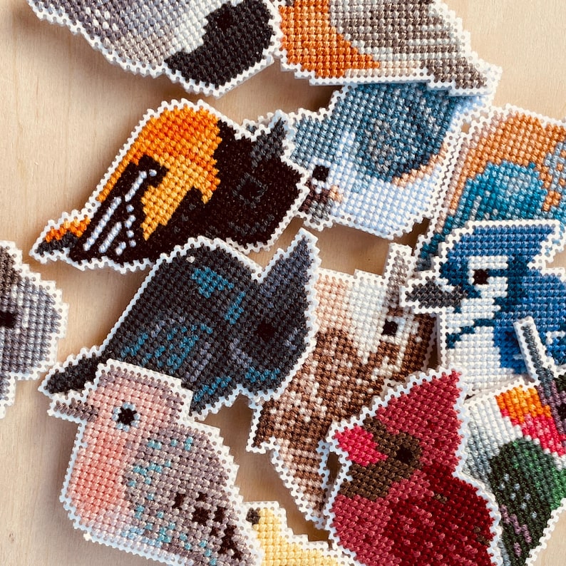 Op de afbeelding: Een verzameling kleurrijke, kruissteek vogelornamenten. De vogels hebben verschillende ontwerpen met tinten blauw, oranje, zwart, rood en bruin. Elke vogel is omlijnd met witte steken, wat een textuur geeft. De ornamenten zijn gerangschikt op een lichtgekleurd houten oppervlak.