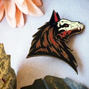 Puede incluir: Pin de esmalte negro y rojo de una cabeza de lobo con un diseño de calavera. El pelaje del lobo es negro con detalles rojos y dorados.