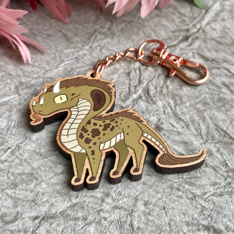 Op de afbeelding: Een houten sleutelhanger met een cartoon groene en bruine draak met een lange nek en staart. De draak heeft bruine vlekken en een witte streep langs zijn nek. De sleutelhanger heeft een ros&eacute;gouden ketting en sluiting.