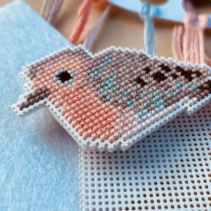 Op de afbeelding: Een close-up van een kruissteek vogelornament in tinten perzik, bruin en blauw. De vogel staat op een wit geperforeerd oppervlak, met draden op de achtergrond. De vogel heeft een klein zwart oog en een witte rand.