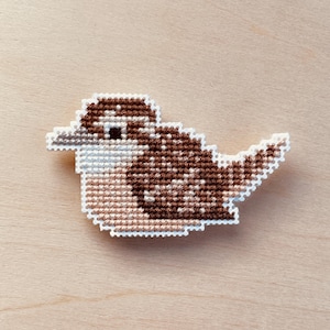 Op de afbeelding: Een kruissteekvogel in bruin-, beige- en wittinten. De vogel is een gedetailleerd, handgemaakt ambacht met een witte omtrek. De vogel staat tegen een lichte houtnerf achtergrond.