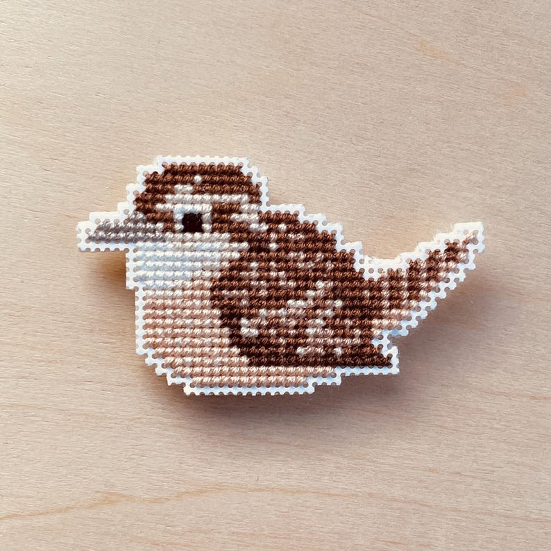 Op de afbeelding: Een kruissteekvogel in bruin-, beige- en wittinten. De vogel is een gedetailleerd, handgemaakt ambacht met een witte omtrek. De vogel staat tegen een lichte houtnerf achtergrond.