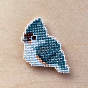 Op de afbeelding: Een kruissteekvogel in tinten blauw, wit, bruin en beige. De vogel is een gestileerd ontwerp met een witte rand. De achtergrond is een lichte houtnerf.