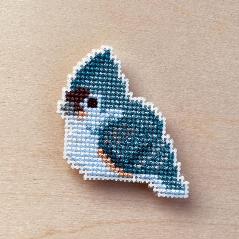 Op de afbeelding: Een kruissteekvogel in tinten blauw, wit, bruin en beige. De vogel is een gestileerd ontwerp met een witte rand. De achtergrond is een lichte houtnerf.