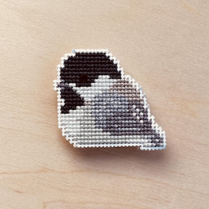 Op de afbeelding: Een handgemaakte kruissteekspeld met een gestileerd vogelontwerp. De vogel is afgebeeld in tinten zwart, grijs en wit, met een witte rand. De speld staat tegen een lichte houtnerf achtergrond.