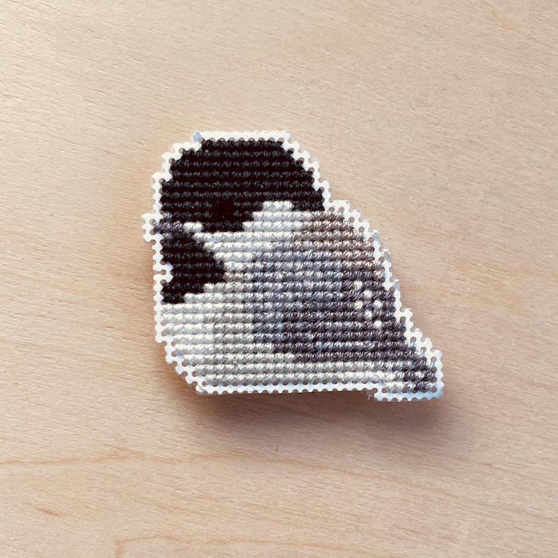 Op de afbeelding: Een handgemaakte kruissteekspeld met een gestileerd vogelontwerp. De vogel is afgebeeld in tinten zwart, grijs en wit, met een witte rand. De speld staat tegen een lichte houtnerf achtergrond.