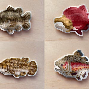 Puede incluir: Cuatro diseños de peces de punto de cruz en tonos marrón, rojo y dorado, cada uno con un borde blanco. Los peces están dispuestos sobre una superficie de madera clara. Los diseños incluyen un bajo, un pez rojo y otros dos peces.