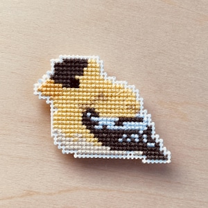 Op de afbeelding: Een kruissteek vogelspeld met een gele body, zwarte kop en blauwe accenten. De vogel is wit omlijnd en geplaatst tegen een lichte houtnerf achtergrond. Deze handgemaakte speld is een uniek accessoire.