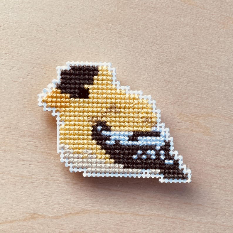 Op de afbeelding: Een kruissteek vogelspeld met een gele body, zwarte kop en blauwe accenten. De vogel is wit omlijnd en geplaatst tegen een lichte houtnerf achtergrond. Deze handgemaakte speld is een uniek accessoire.