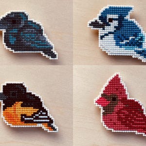May include: Four cross-stitch bird designs: a dark blue raven, a blue jay, an orange and black oriole, and a red cardinal. Each bird is outlined in white and set against a light wood grain background.