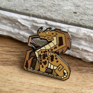 Op de afbeelding: Een bruine en beige emaille pin van een gestileerde draak met een geel oog. De draak is opgerold met zijn staart onder zijn lichaam.