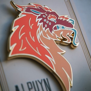 May include: A gold-toned enamel pin featuring a red, orange, and blue stylized wolf head with a white outline. The pin is on a white background with the word "ALPUYN" printed below it.