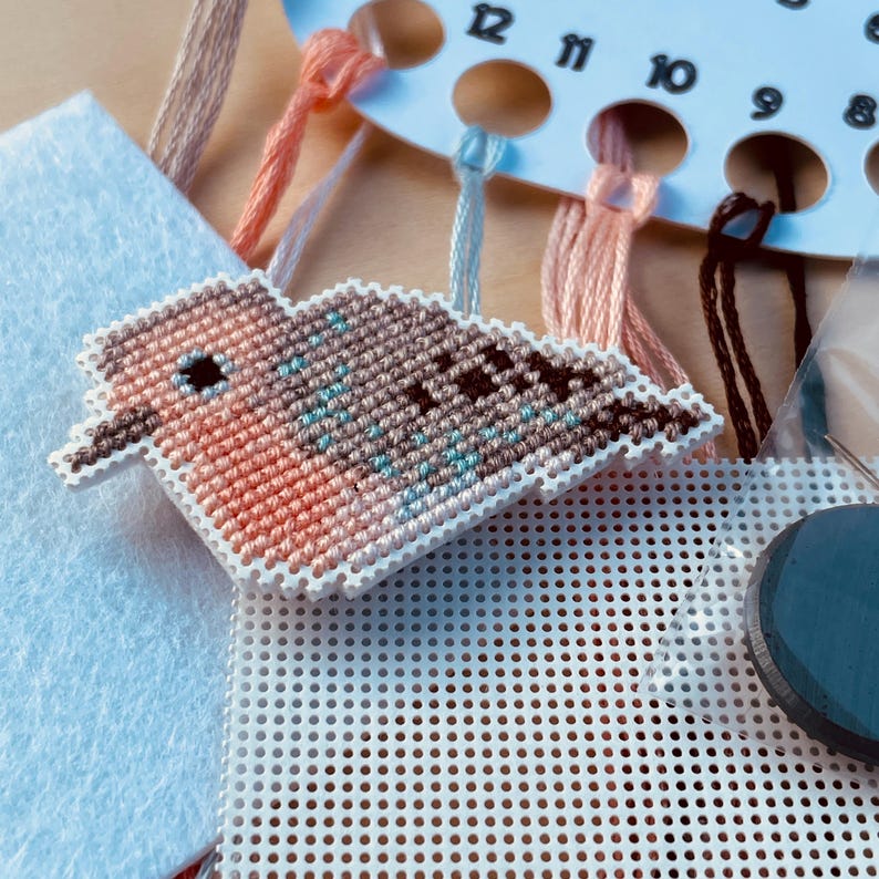 Op de afbeelding: Een kruissteekpakket met een vogelontwerp in bruin, perzik en blauw. De set bevat een wit plastic canvas, borduurgaren, een naald en een vierkant vilt. De vogel is het middelpunt, met gedetailleerde steken.