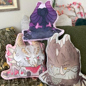 May include: Three plush pillows featuring different monster designs. The first pillow is purple and black with a monster with horns and a large mouth. The second pillow is pink and white with a monster with a long snout and red eyes. The third pillow is brown and white with a monster with a long snout and yellow eyes.