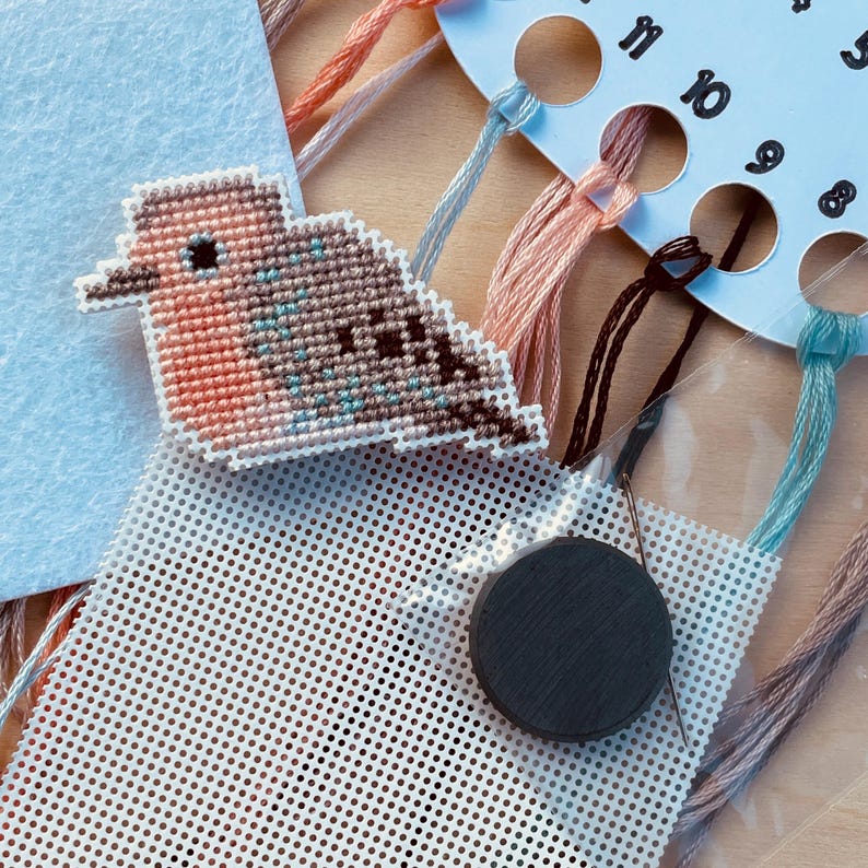 Op de afbeelding: Een kruissteekset met een vogelontwerp. De set bevat een geborduurde vogel, borduurgaren in verschillende kleuren, een naald, een ronde magneet en een geperforeerd plastic canvas. De vogel is voornamelijk perzik, bruin en blauw.