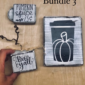 May include: Three wooden signs with fall-themed sayings. The first sign says "Pumpkin Spice $2.25", the second sign says "Boo Y'all", and the third sign has a black silhouette of a coffee cup with a pumpkin inside.