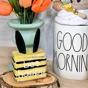 May include: A wooden block painted yellow with black stripes and the words "bee happy" in white. The block is sitting on a wooden slice and has black bunny ears.