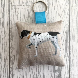 May include: A square beige fabric keyring with a painted illustration of a dog with black spots and a brown collar. It has a silver ring and a blue fabric loop. The dog is shown in a walking pose. The keyring is a decorative item.